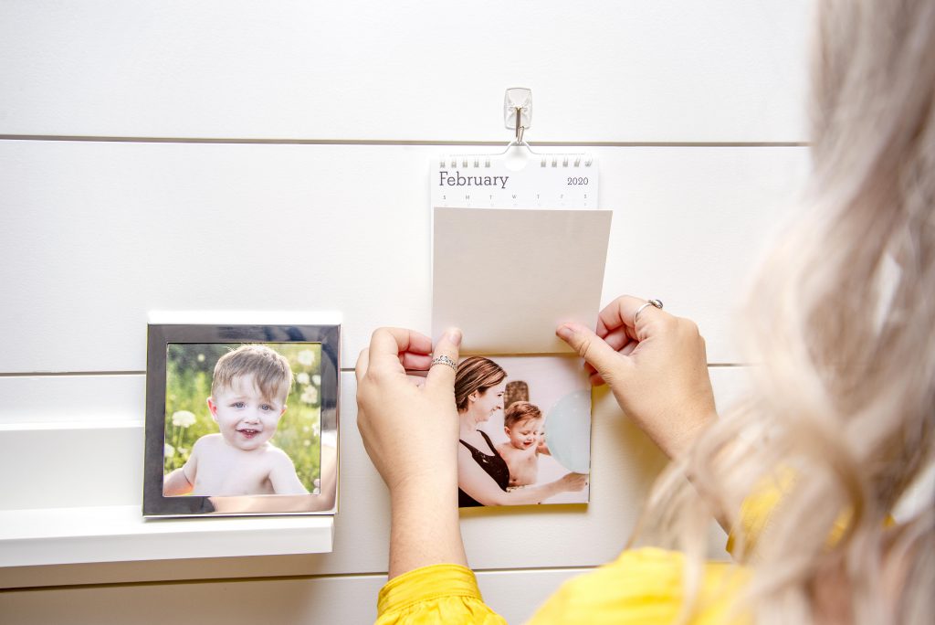 Adult blonde female shown holding the page of a hanging wall photo calendar. Silver frame with the photo of a baby boy shown on a shelf next to the calendar.