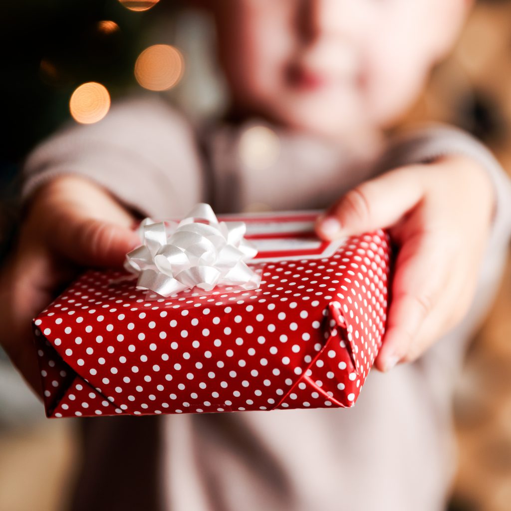 blurred outline of small boy, in focus are his two hands holding small box wrapped in red paper with white polka dots and white bow